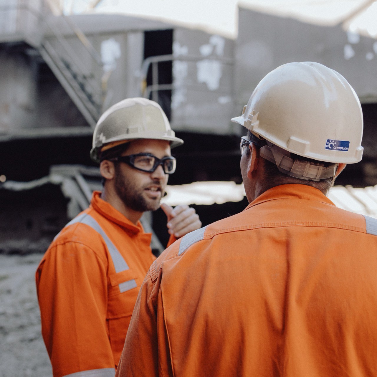 Twee procesoperators in oranje veiligheidskleding en witte helmen bespreken werkzaamheden op een industriële locatie.